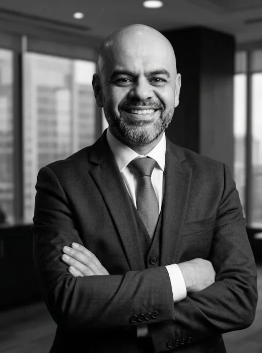 Professional headshot of a bald man in a dark suit and tie, smiling with arms crossed in a modern office setting
