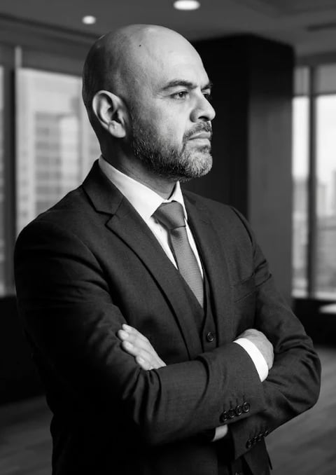 Bald man with beard in dark suit and tie standing with arms crossed in modern office setting, black and white portrait
